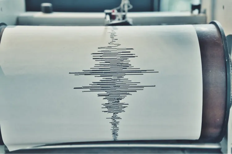 A close-up of a drum seismograph with its needle tracing a jagged black earthquake waveform onto a roll of white graph paper.
