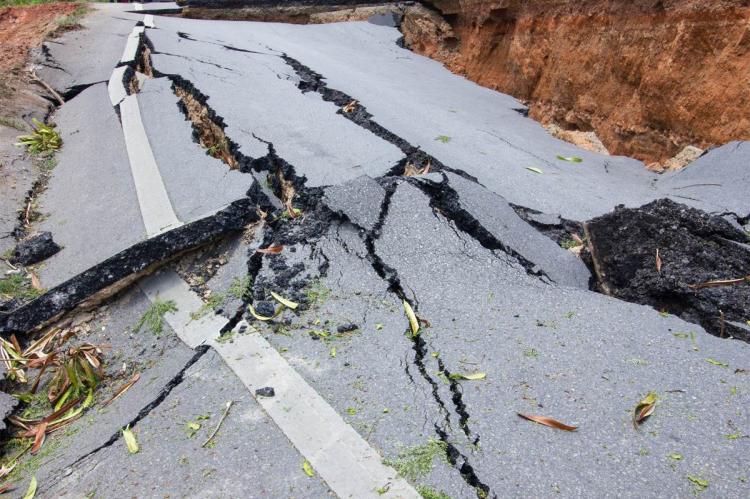 A tarmac road torn apart by an earthquake, its surface split into large slabs by deep cracks and dropped away into a red-earth ravine at one side.