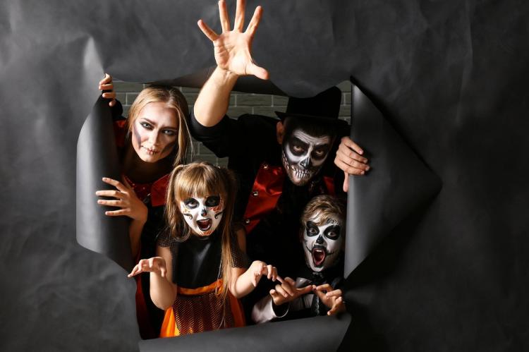 A family of four in Halloween costumes and skeleton-style face paint — two parents and two children — peering and reaching through a ragged hole torn in a grey paper backdrop, pulling spooky faces.