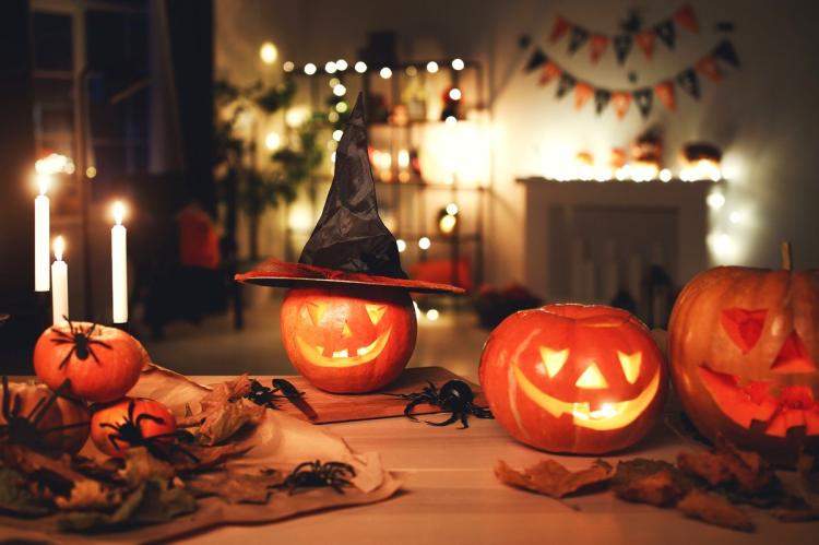 Three carved jack-o'-lanterns glowing on a wooden table — the central one wearing a black witch's hat — surrounded by lit candles, plastic spiders and autumn leaves, with string lights and bunting on shelves behind.