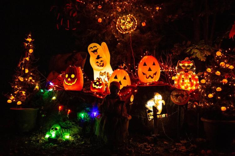 A night-time garden Halloween display with glowing pumpkin, ghost and cat lanterns, strings of fairy lights, a luminous green skull and a child in costume silhouetted in the centre.
