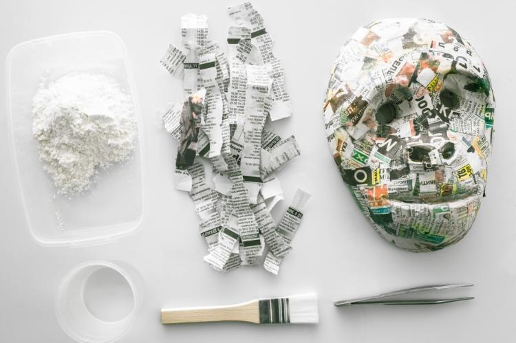 Flat lay of papier-mâché supplies on a white surface: a tub of flour paste, torn strips of newspaper, a finished newspaper mask, a water pot, a brush and tweezers.