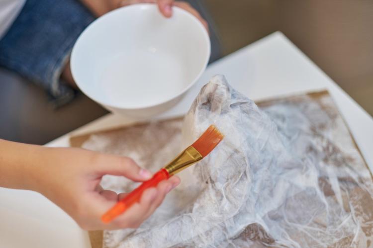 A child brushing paste over a plastic-wrapped form on a lined tray, shaping a papier-mâché mountain, with a white bowl held above.