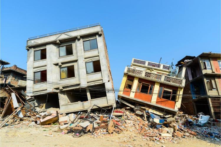 Two multi-storey concrete buildings left leaning and partly collapsed after an earthquake, surrounded by piles of brick and concrete rubble under a clear blue sky.