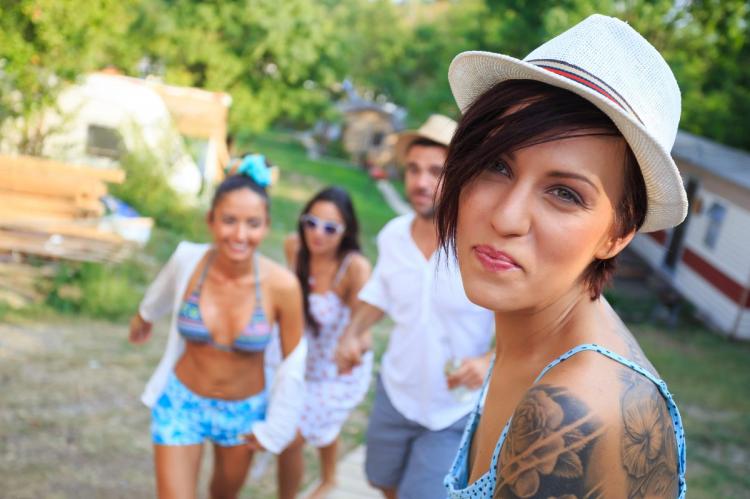 A woman with a white fedora and a shoulder tattoo looks at the camera, with three friends in summery attire in the blurred background.